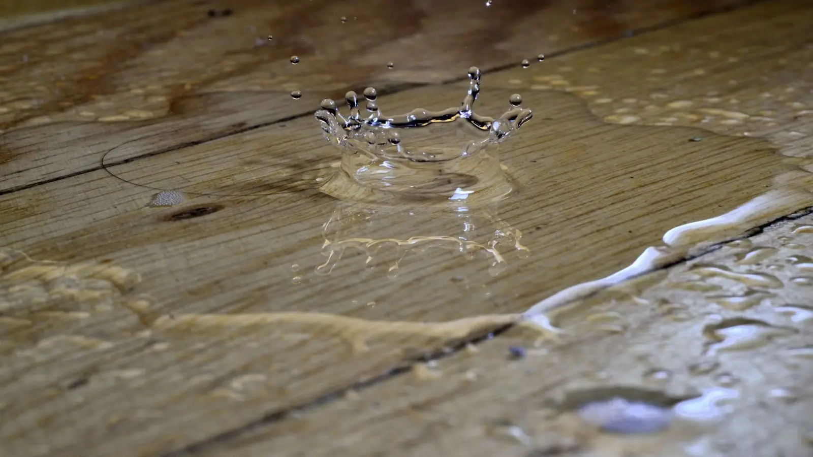 Water splashing on a wet hardwood plank floor with moisture pooling along the seams