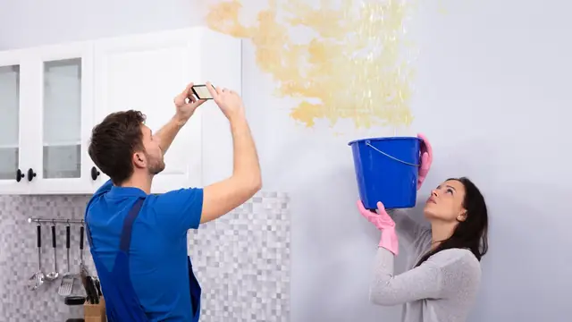 Man photographing a water-stained wall while a woman holds a bucket to catch a leak
