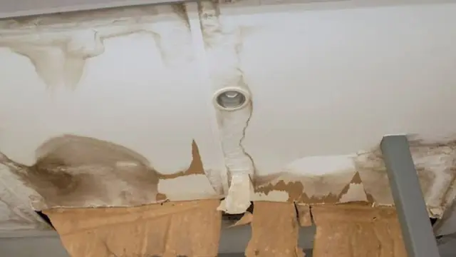 Water-stained ceiling with brown patches, peeling paint and sagging material around a pipe opening